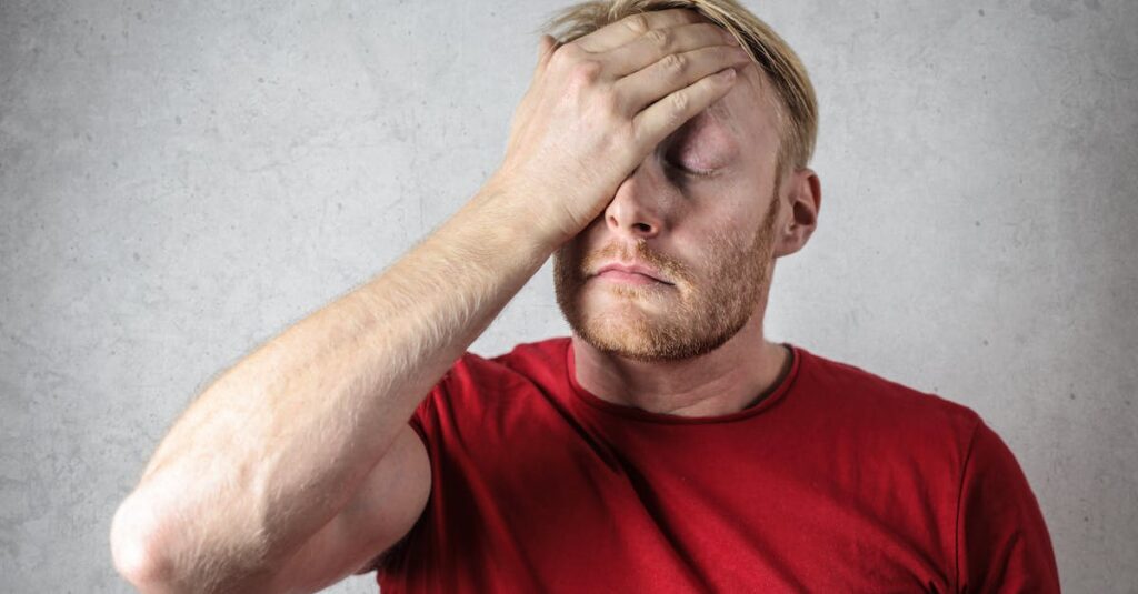 A frustrated man in a red shirt holds his head in stress against a neutral background.