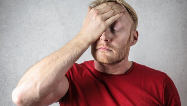 A frustrated man in a red shirt holds his head in stress against a neutral background.