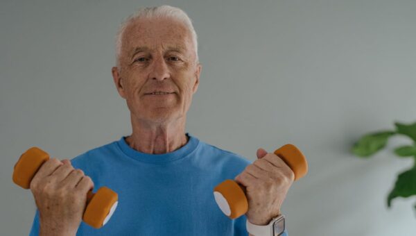 Elderly man in blue shirt lifting dumbbells and smiling indoors, promoting active lifestyle.