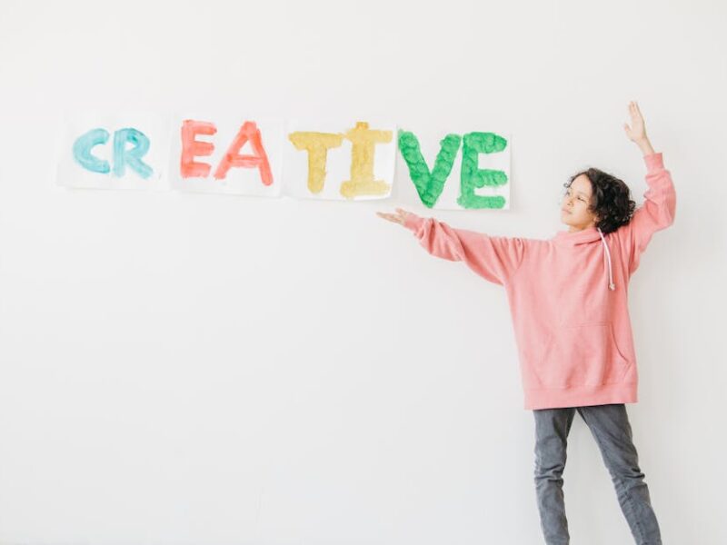 A child with curly hair in a pink hoodie points to colorful 'CREATIVE' text on a white wall.