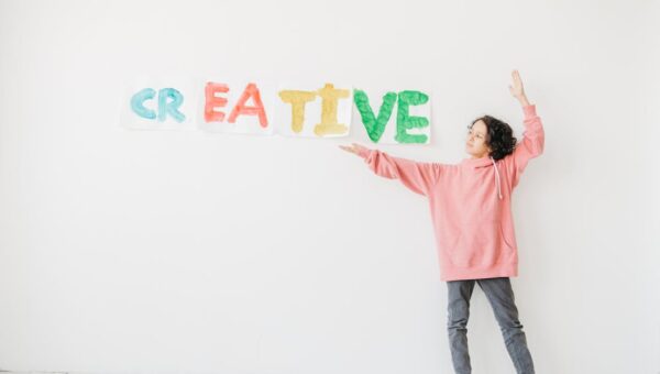 A child with curly hair in a pink hoodie points to colorful 'CREATIVE' text on a white wall.