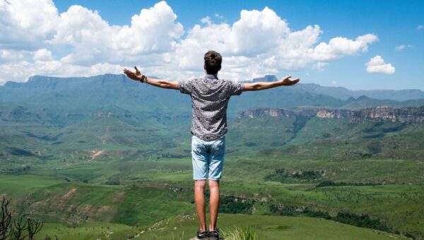 A man stands triumphantly with arms open, overlooking a scenic mountain range.