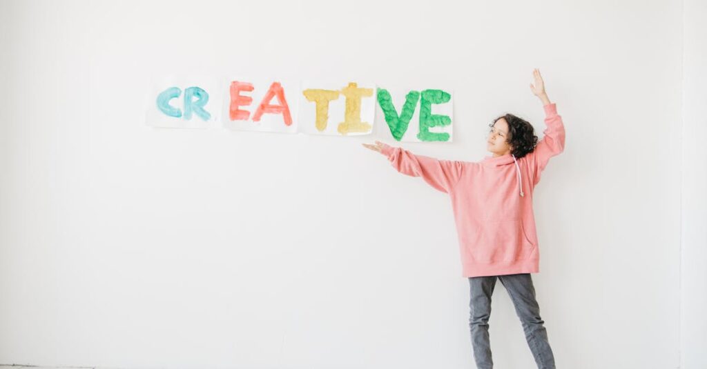 5 Maneiras de Cultivar a Criatividade no Dia a Dia 2 A child with curly hair in a pink hoodie points to colorful 'CREATIVE' text on a white wall.