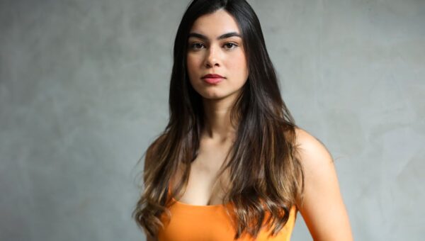 Elegant portrait of a young woman with long, dark hair in a vibrant orange top against a neutral background.
