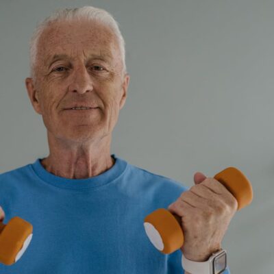 Elderly man in blue shirt lifting dumbbells and smiling indoors, promoting active lifestyle.