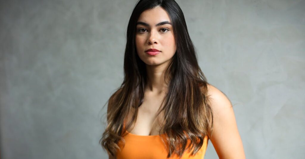 Elegant portrait of a young woman with long, dark hair in a vibrant orange top against a neutral background.