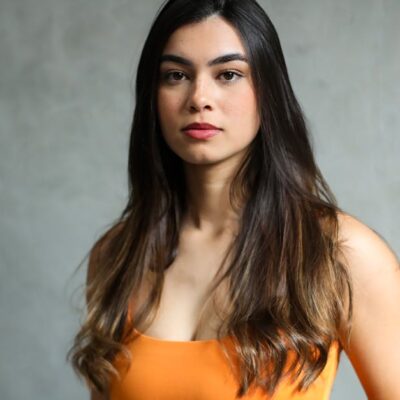 Elegant portrait of a young woman with long, dark hair in a vibrant orange top against a neutral background.