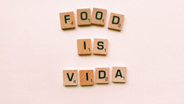 Wooden letter tiles spell 'FOOD IS VIDA' on a soft pink background.