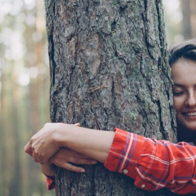 Smiling woman embracing a tree in a sunlit forest, promoting nature connection.