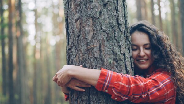 Smiling woman embracing a tree in a sunlit forest, promoting nature connection.