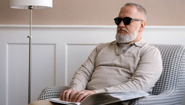 Senior man with sunglasses reading a braille book in a cozy modern interior.