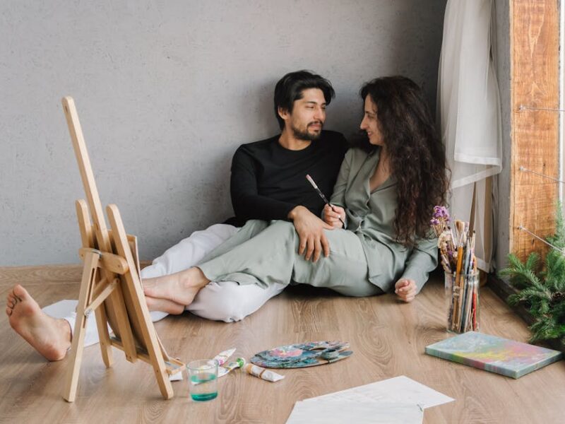 Happy couple sitting on the floor inside, enjoying a painting session with natural light.