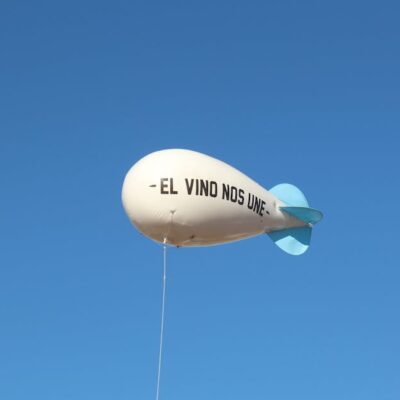 A white balloon with 'El Vino Nos Une' phrase floats against a clear blue sky.