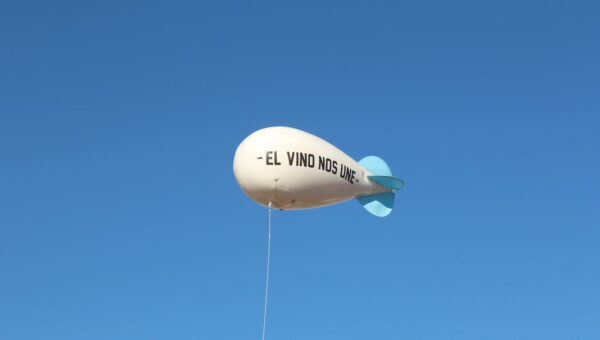 A white balloon with 'El Vino Nos Une' phrase floats against a clear blue sky.