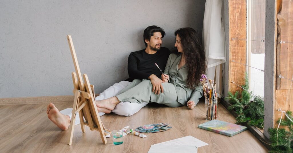 Happy couple sitting on the floor inside, enjoying a painting session with natural light.
