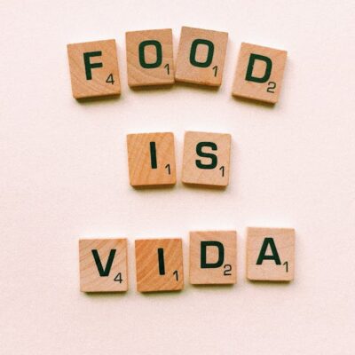 Wooden letter tiles spell 'FOOD IS VIDA' on a soft pink background.