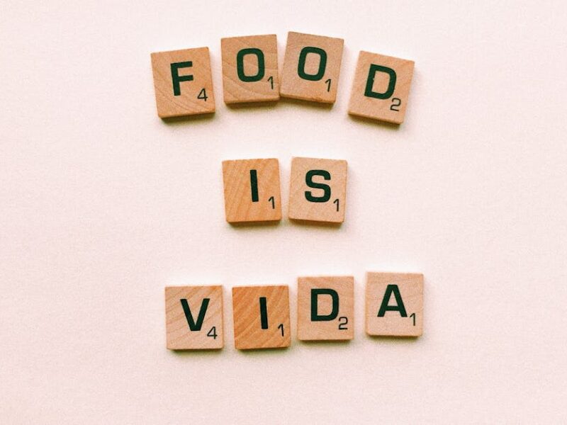 Wooden letter tiles spell 'FOOD IS VIDA' on a soft pink background.