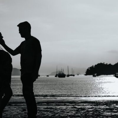 Black and white full body side view of anonymous couple caressing on shore near sea with various boats against pier