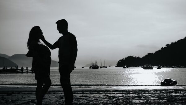 Black and white full body side view of anonymous couple caressing on shore near sea with various boats against pier