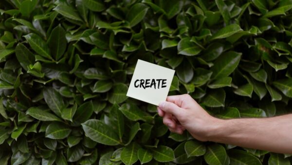 A hand holding a note with 'Create' written on it against a lush green leafy background.