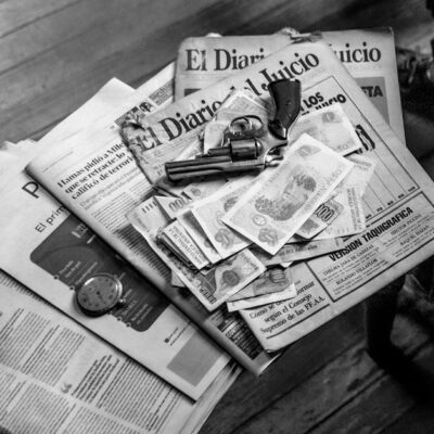 Black and white image featuring currency, newspapers, and a revolver on a wooden surface.