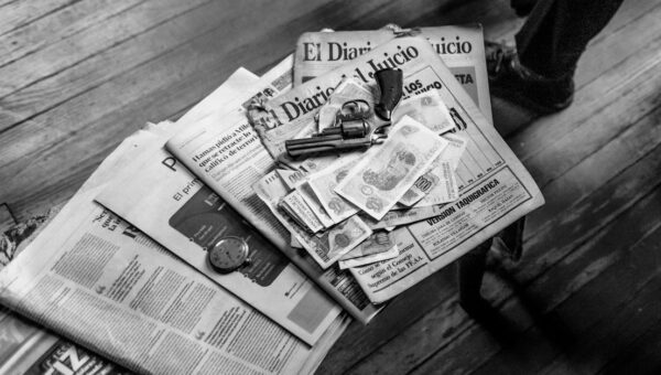 Black and white image featuring currency, newspapers, and a revolver on a wooden surface.