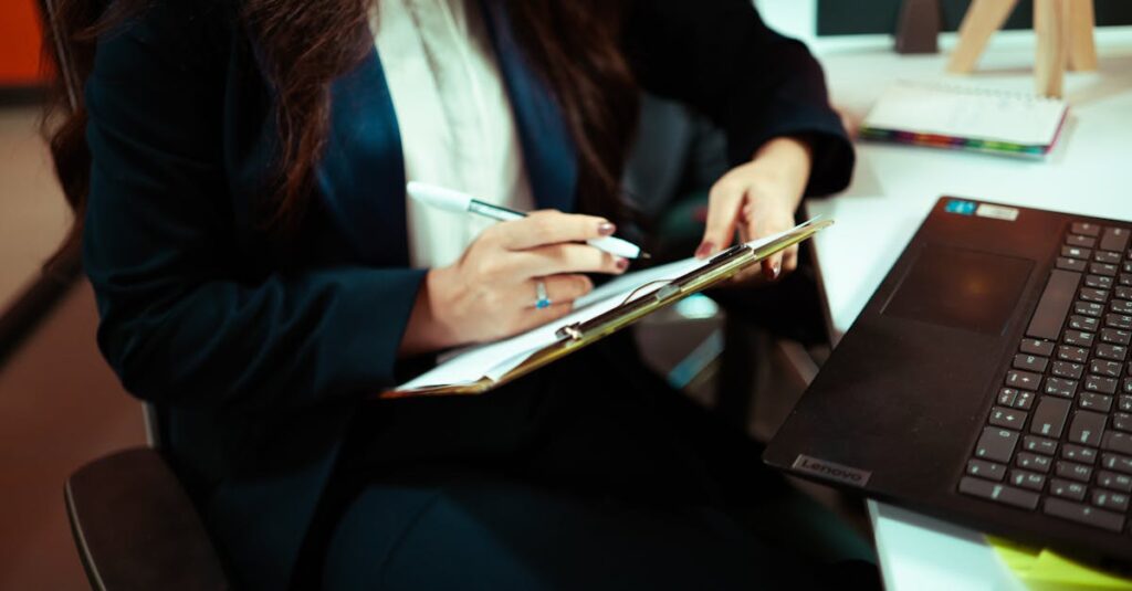 A focused businesswoman writing on a clipboard with a laptop nearby in an office environment.