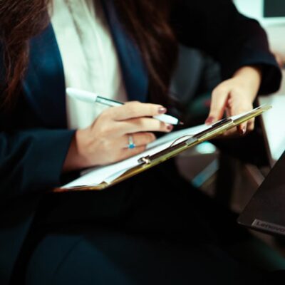 A focused businesswoman writing on a clipboard with a laptop nearby in an office environment.