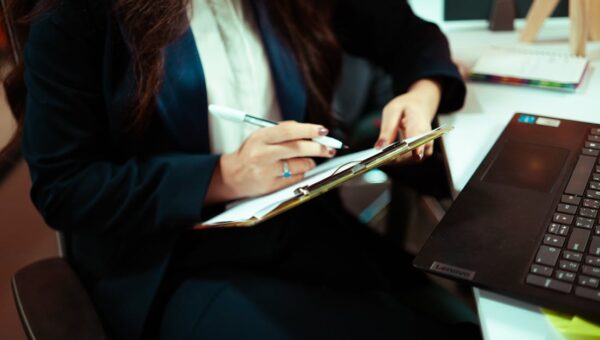 A focused businesswoman writing on a clipboard with a laptop nearby in an office environment.