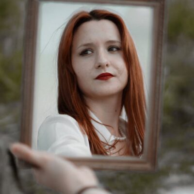 A young redhead woman holding a mirror outdoors, showcasing her reflection with red lips.