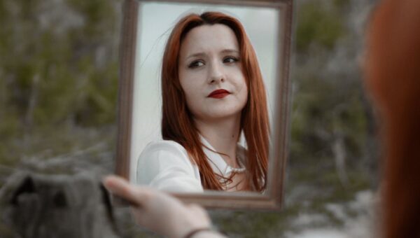 A young redhead woman holding a mirror outdoors, showcasing her reflection with red lips.