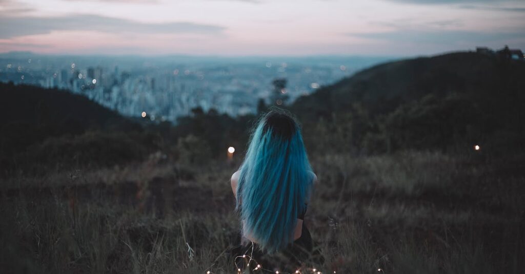 A woman with blue hair sits on a hillside, gazing at the city's twilight skyline adorned with string lights.