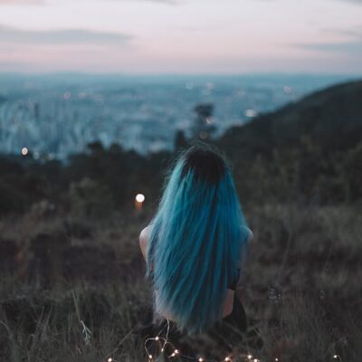 A woman with blue hair sits on a hillside, gazing at the city's twilight skyline adorned with string lights.