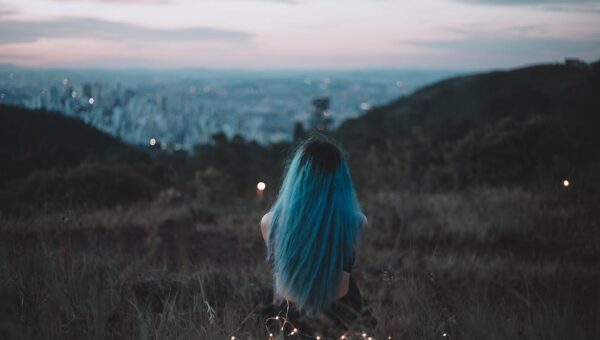 A woman with blue hair sits on a hillside, gazing at the city's twilight skyline adorned with string lights.