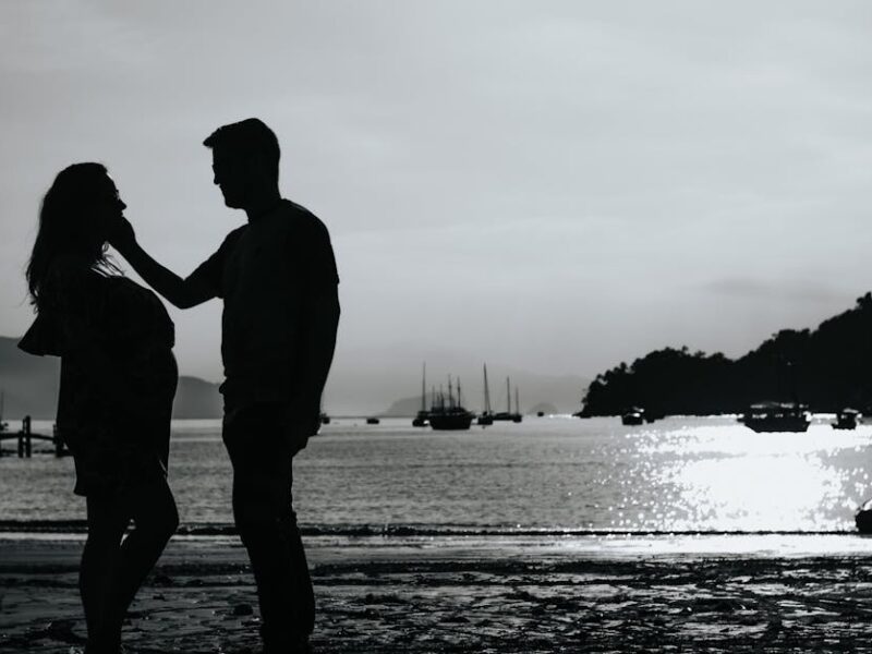 Black and white full body side view of anonymous couple caressing on shore near sea with various boats against pier