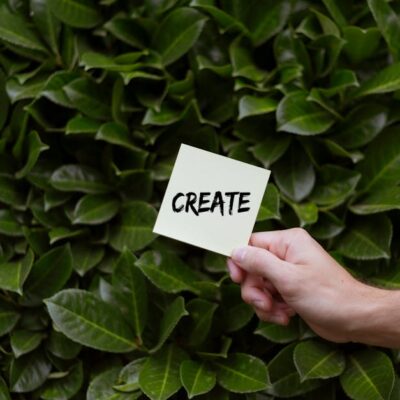A hand holding a note with 'Create' written on it against a lush green leafy background.