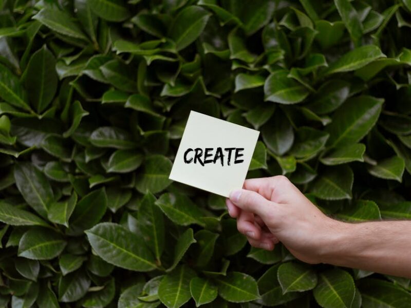 A hand holding a note with 'Create' written on it against a lush green leafy background.