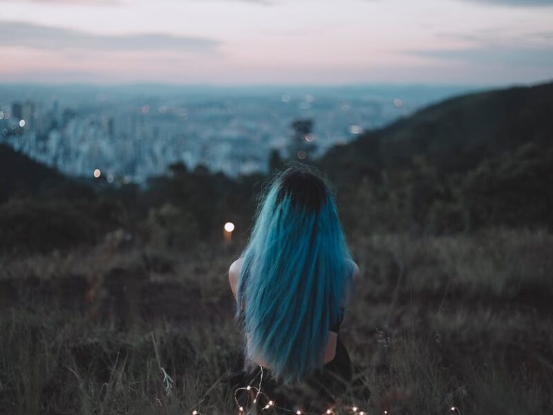 A woman with blue hair sits on a hillside, gazing at the city's twilight skyline adorned with string lights.