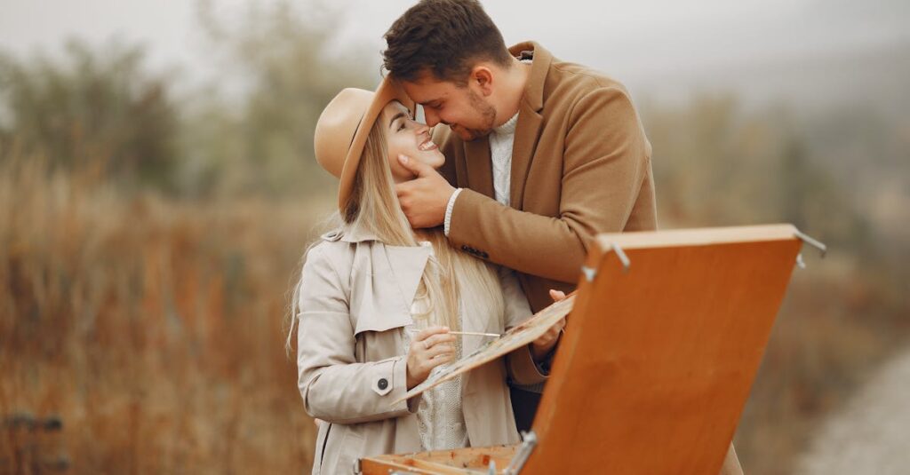 Young happy woman in hat with palette and man in trendy coat smiling and hugging against portable easel in park