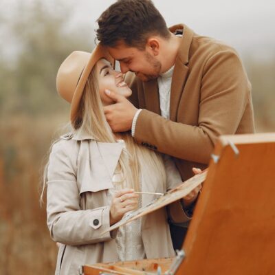 Young happy woman in hat with palette and man in trendy coat smiling and hugging against portable easel in park