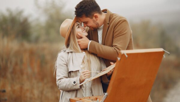 Young happy woman in hat with palette and man in trendy coat smiling and hugging against portable easel in park