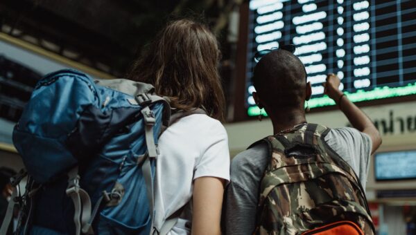 Two backpackers checking the digital board at a train station for travel information.