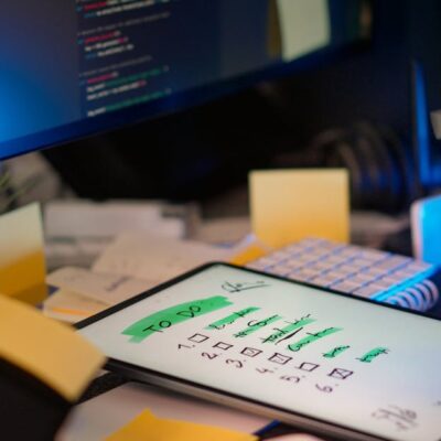 A cluttered desk with a tablet showing a to-do list, monitor showing code, and office supplies.