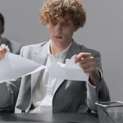 Two employees in suits sitting at desks, one tearing paper.