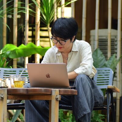 A woman focuses on her laptop while sitting in a lush outdoor cafe.