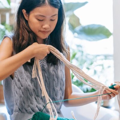 Asian woman skillfully crocheting with yarn in a comfortable indoor space surrounded by plants.