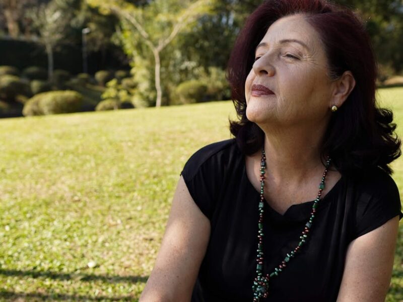 A relaxed, mature woman enjoys the sunlight in a tranquil park setting, exuding peace and contentment.