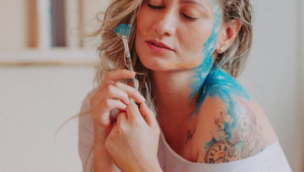 An artistic portrait of a woman with face and body paint holding a brush, eyes closed.