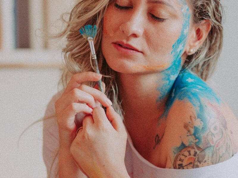 An artistic portrait of a woman with face and body paint holding a brush, eyes closed.