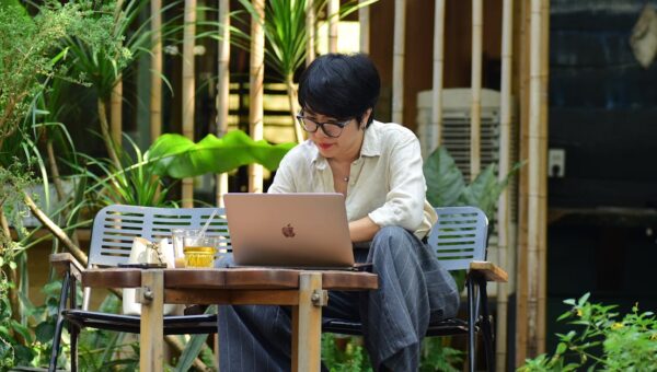 A woman focuses on her laptop while sitting in a lush outdoor cafe.